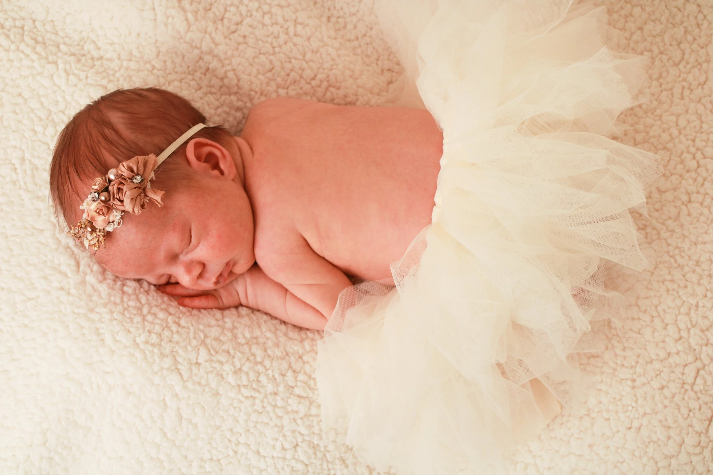 Newborn baby girl wearing an ivory tutu and floral headband, sleeping on a cream blanket during a styled newborn session
